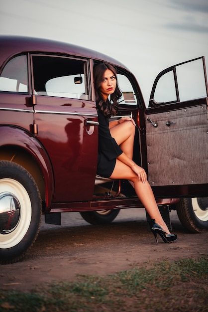 Photo jeune fille brune sexy avec de longues jambes dans une robe noire et des chaussures à talons hauts sort d'une voiture rétro bordeaux sur une route de campagne