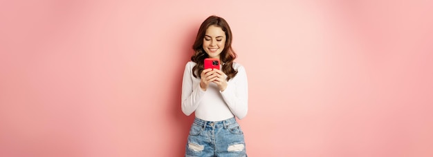 Jeune fille brune souriante à l'aide d'une commande de message SMS sur smartphone en ligne sur l'application de téléphone mobile debout sur fond rose
