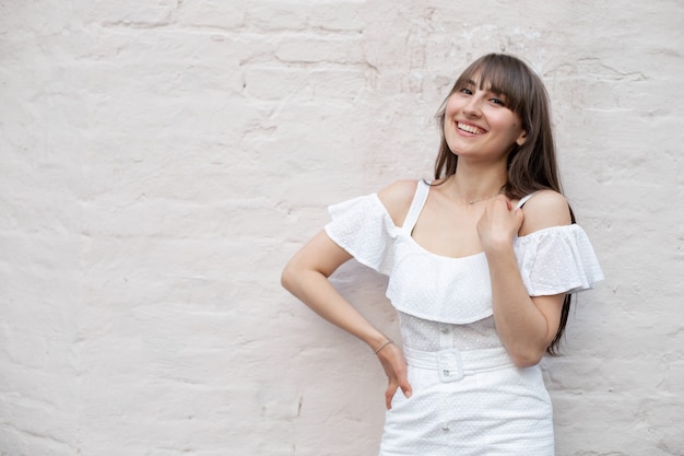 Jeune fille brune souriante aux cheveux longs dans une robe blanche sur un fond de brique solide. place pour votre conception