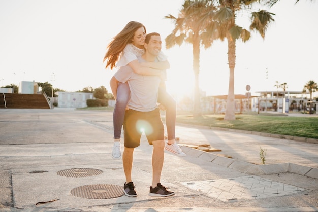 Une jeune fille brune souriante chevauche son petit ami hispanique entre les palmiers et les rayons du soleil en Espagne