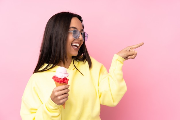 Jeune fille brune tenant une glace cornet sur fond rose isolé doigt pointé sur le côté et présentant un produit