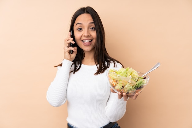 Jeune fille brune tenant une salade sur un mur isolé en gardant une conversation avec le téléphone mobile avec quelqu'un