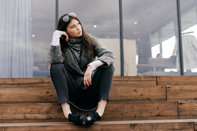 Jeune fille brune urbaine en manteau gris à la mode est assise sur des marches en plein air, pense à quelque chose