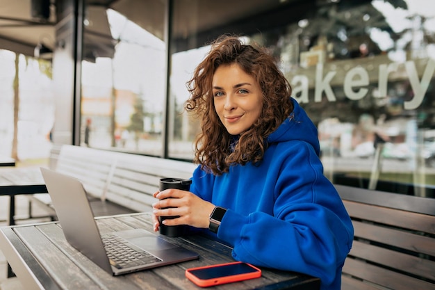 Jeune fille caucasienne animée buvant du café le matin et utilisant son ordinateur portable Une femme brune en vêtements décontractés est assise sur la terrasse d'été Concept de temps libre