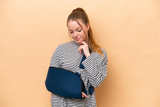 Jeune fille caucasienne avec un bras cassé et portant une écharpe isolée sur fond beige avec une expression triste