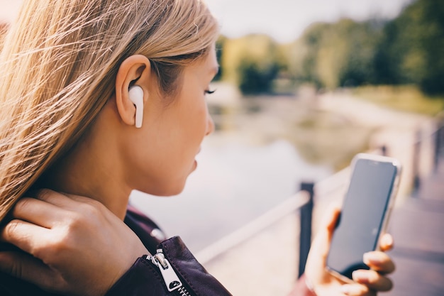 Jeune fille caucasienne avec un casque sans fil dans le parc à l'aide d'un téléphone tablette et souriant