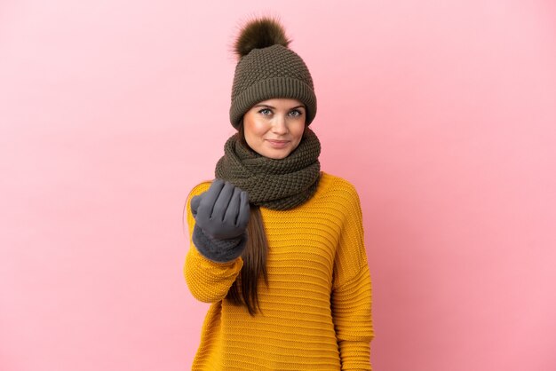 Jeune fille caucasienne avec chapeau d'hiver isolé sur fond rose invitant à venir avec la main. Heureux que tu sois venu