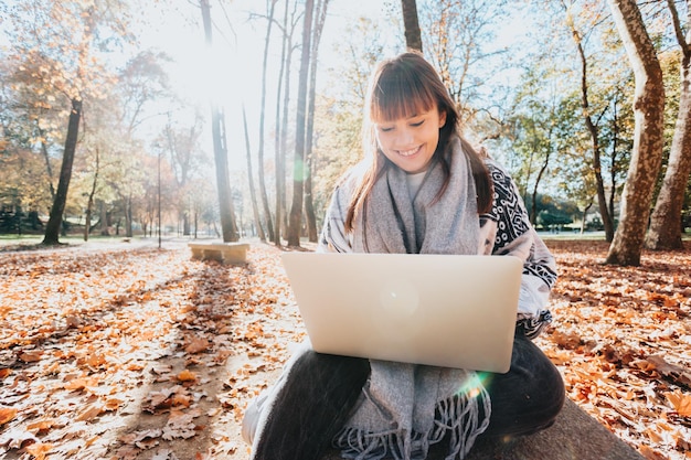 Jeune fille caucasienne streamer à l'extérieur à l'aide d'un ordinateur portable pour vérifier le travail, attitude heureuse et souriante. Style tendance jeune et moderne. Saison d'automne. Travailler n'importe où, liberté financière, investissements