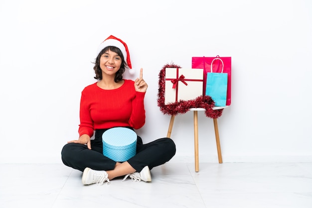 Jeune fille célébrant Noël assis sur le sol isolé sur fond blanc montrant et levant un doigt en signe du meilleur