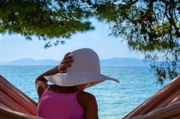 Jeune fille avec un chapeau de paille blanc assis dans un hamac sous le pin surplombant une plage