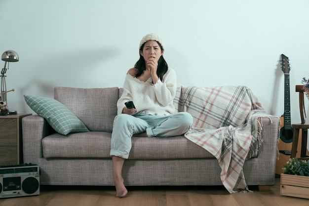 Jeune fille charmante asiatique en chandail regardant un film d'horreur tenant une télévision télécommandée dans le salon de la maison par un mur blanc. femme choquée surprise et effrayée sur le canapé. concept de divertissement intérieur.