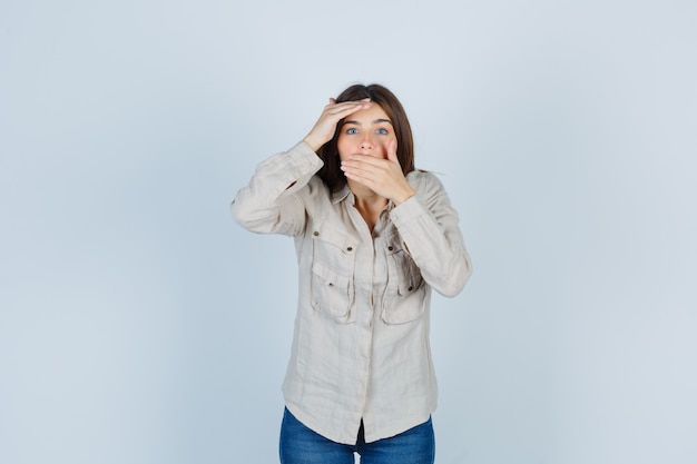 Jeune fille en chemise beige, jeans couvrant la bouche avec la main, regardant au loin avec la main sur la tête et l'air surpris, vue de face.