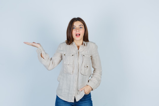 Jeune fille en chemise beige, jeans écartant la paume de la main, la main sur la chemise et l'air surpris, vue de face.