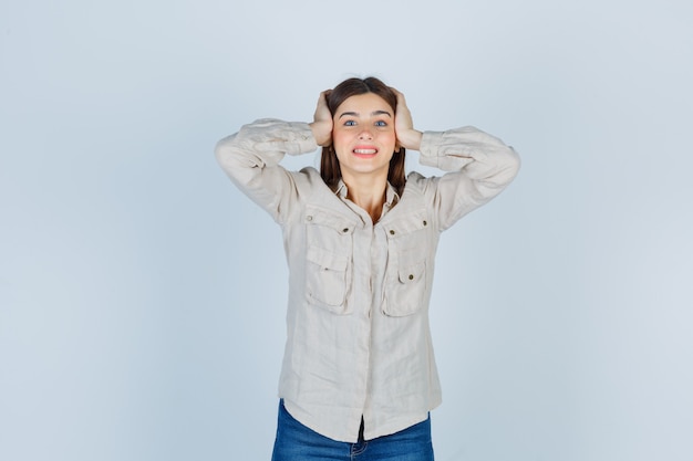 Jeune fille en chemise beige, jeans pressant les mains sur les oreilles, souriante et souriante, vue de face.