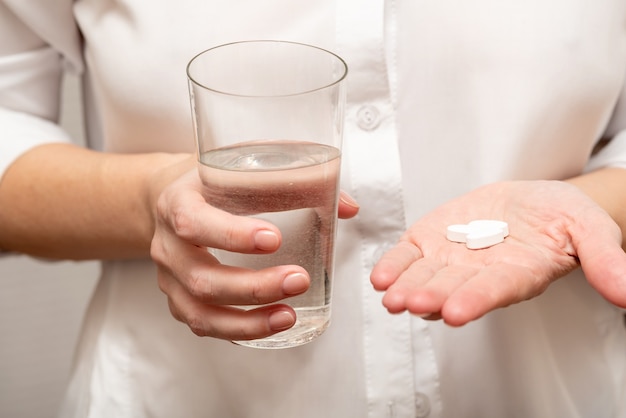 Une jeune fille en chemise blanche tient des vitamines et un verre d'eau dans ses mains.