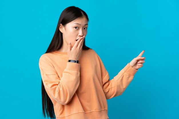 Jeune fille chinoise sur bleu isolé avec une expression de surprise tout en pointant le côté