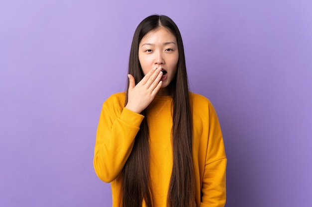 Jeune fille chinoise sur fond violet isolé bâillant et couvrant la bouche grande ouverte avec la main