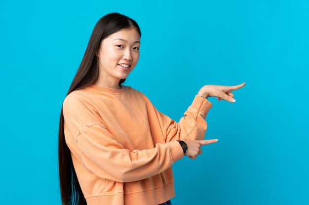 Jeune fille chinoise sur mur bleu isolé pointant le doigt sur le côté et présentant un produit