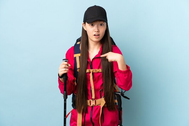 Jeune fille chinoise avec sac à dos et bâtons de randonnée sur bleu isolé pointant vers soi