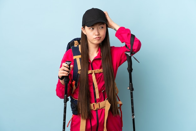 Jeune fille chinoise avec sac à dos et bâtons de randonnée sur mur bleu isolé ayant des doutes tout en se grattant la tête
