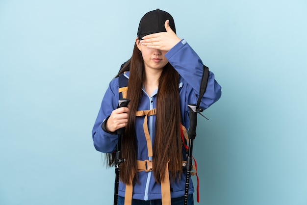 Jeune fille chinoise avec sac à dos et bâtons de randonnée sur mur bleu isolé couvrant les yeux par les mains. Je ne veux pas voir quelque chose