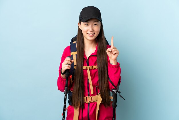 Jeune fille chinoise avec sac à dos et bâtons de randonnée sur un mur bleu isolé pointant vers le haut une excellente idée