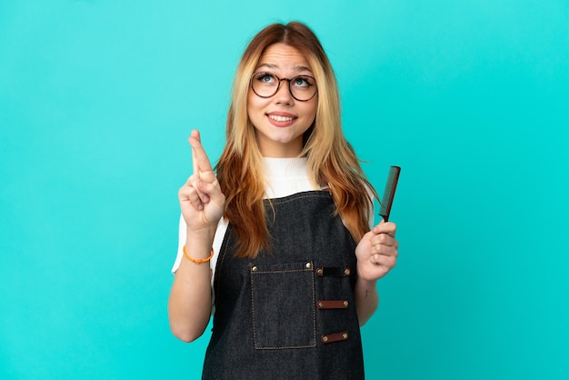 Jeune fille de coiffeur sur fond bleu isolé avec les doigts croisés et souhaitant le meilleur