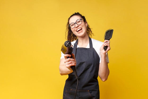 Jeune fille coiffeuse en uniforme tient un sèche-cheveux et un peigne et sourit sur fond jaune