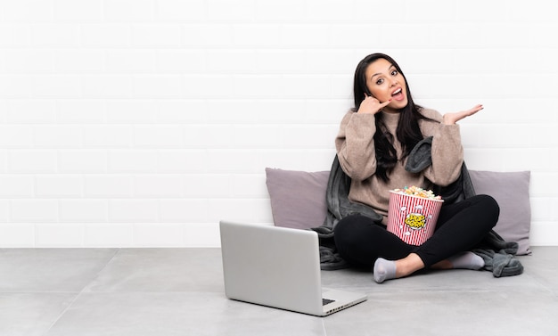 Jeune fille colombienne tenant un bol de pop-corn et montrant un film dans un ordinateur portable faisant un geste de téléphone et doutant