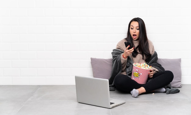 Jeune fille colombienne tenant un bol de pop-corn et montrant un film dans un ordinateur portable surpris et envoyant un message