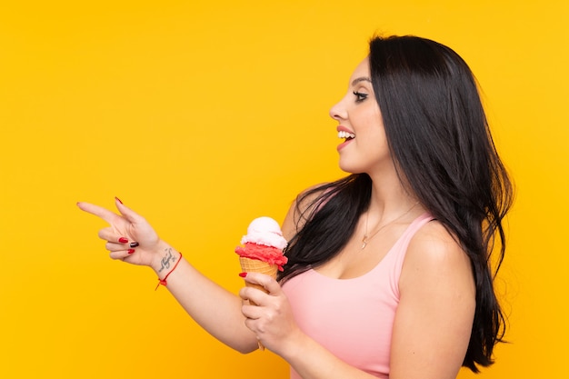 Jeune fille colombienne tenant une glace au cornet sur un mur jaune pointant vers le côté pour présenter un produit