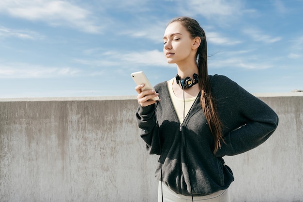 Une jeune fille confiante en vêtements de sport noirs est allée à une séance d'entraînement en plein air regarde dans son smartphone