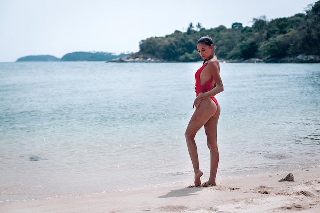 Jeune fille avec un corps magnifique se repose sur la plage de sable blanc près de l'océan. Beau modèle sexy dans un bain de soleil maillot de bain rouge. Brune aux cheveux bouclés en bikini.