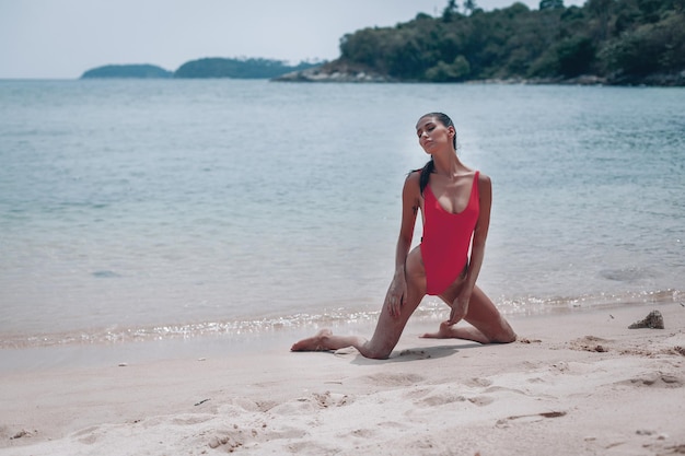 Jeune fille avec un corps magnifique se repose sur la plage de sable blanc près de l'océan. Beau modèle sexy dans un bain de soleil maillot de bain rouge. Brune aux cheveux bouclés en bikini.