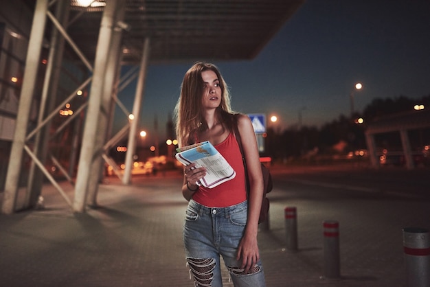 Jeune fille coûte la nuit près du terminal de l'aéroport ou de la gare et lit le plan de la ville et cherche un hôtel. Le touriste mignon avec des sacs à dos détermine le concept de voyage.