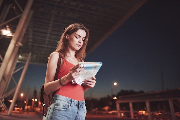 Jeune fille coûte la nuit près du terminal de l'aéroport ou de la gare et lit le plan de la ville et cherche un hôtel. Le touriste mignon avec des sacs à dos détermine le concept de voyage.