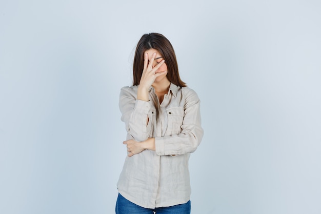 Jeune fille couvrant le visage avec la main en chemise beige, jeans et l'air ennuyé, vue de face.