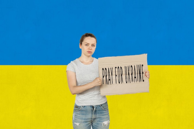 Jeune fille criant tenant une affiche avec lettrage Priez pour l'Ukraine isolée sur fond de drapeau bleu et jaune ukrainien