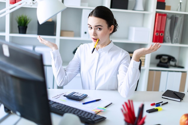 Une jeune fille dans le bureau est assise à une table, tenant un stylo dans la bouche et écartant les mains.