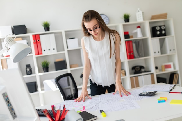 Une jeune fille dans le bureau est debout près de la table et regarde des documents.