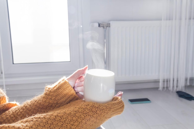 Une jeune fille dans un chandail jaune et un chapeau boit du thé chaud près d'un radiateur avec un thermostat