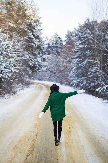 Une jeune fille dans un chandail vert et un chapeau marche au milieu d'une route enneigée dans une épaisse forêt de pins Jour de gel