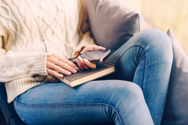 Jeune fille dans un pull blanc et un jean assis sur le rebord de la fenêtre tenant un téléphone dans les mains et écouter de la musique