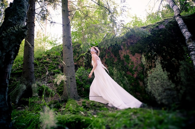 Une jeune fille dans une robe blanche et un bandeau dans une forêt au rocher en mousse