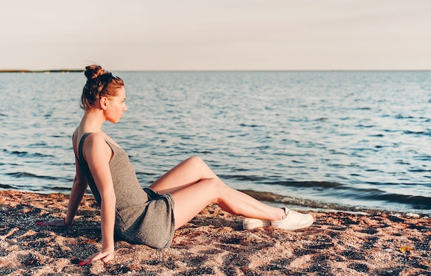 jeune fille dans une robe légère et des lunettes de soleil assis sur le sable et regardant la mer