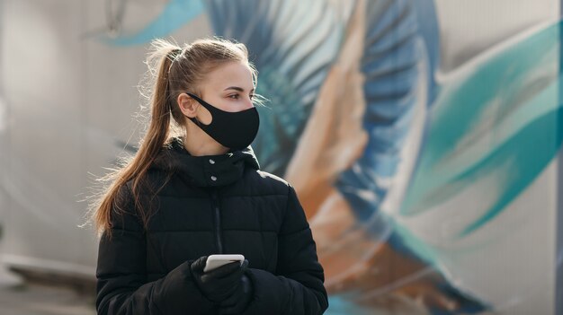 Jeune fille dans la rue de la ville portant un masque facial médical stérile noir. Femme utilisant le téléphone pour rechercher des nouvelles sur nCov 2019. Quarantaine COVID-19 pandémie de coronavirus pandémique et concept de soins de santé