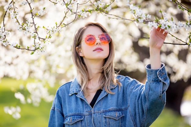 Jeune fille dans une veste en jean et lunettes de soleil se tient près d&#39;un arbre en fleurs