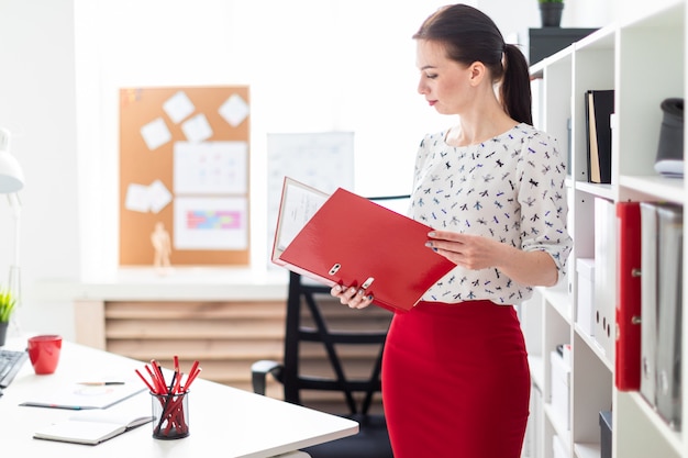 Une jeune fille debout dans le bureau et tenant un dossier rouge avec des documents.