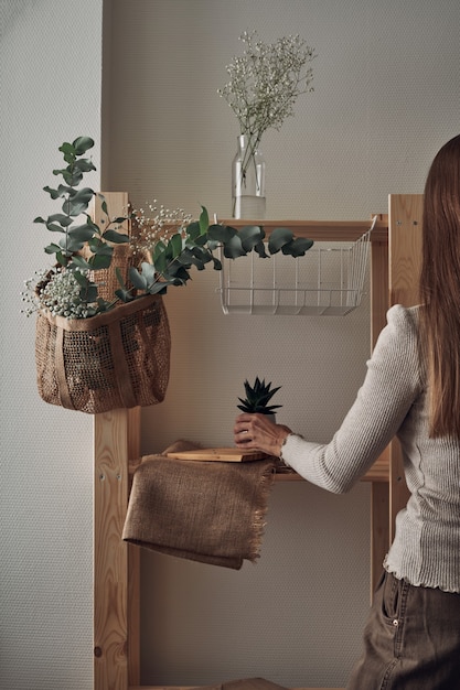 La jeune fille décore un support en bois avec un sac écologique avec des branches d'eucalyptus, des fleurs, des plantes d'intérieur