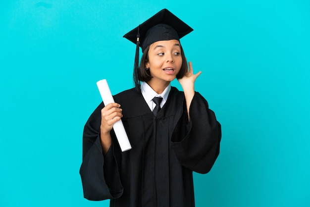 Jeune fille diplômée de l'université sur fond bleu isolé écoutant quelque chose en mettant la main sur l'oreille